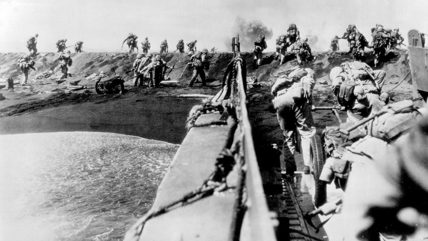 Fourth Marines Hit Iwo Jima Beach -- Fourth Marines dash from landing craft, dragging equipment, while others Go Over The Top of sand dune as they hit the beach of Iwo Jima, Volcano Islands, February 19. Smoke of artillery of Mortar fire in background. February 22, 1945. (Photo by Joe Rosenthal, AP).