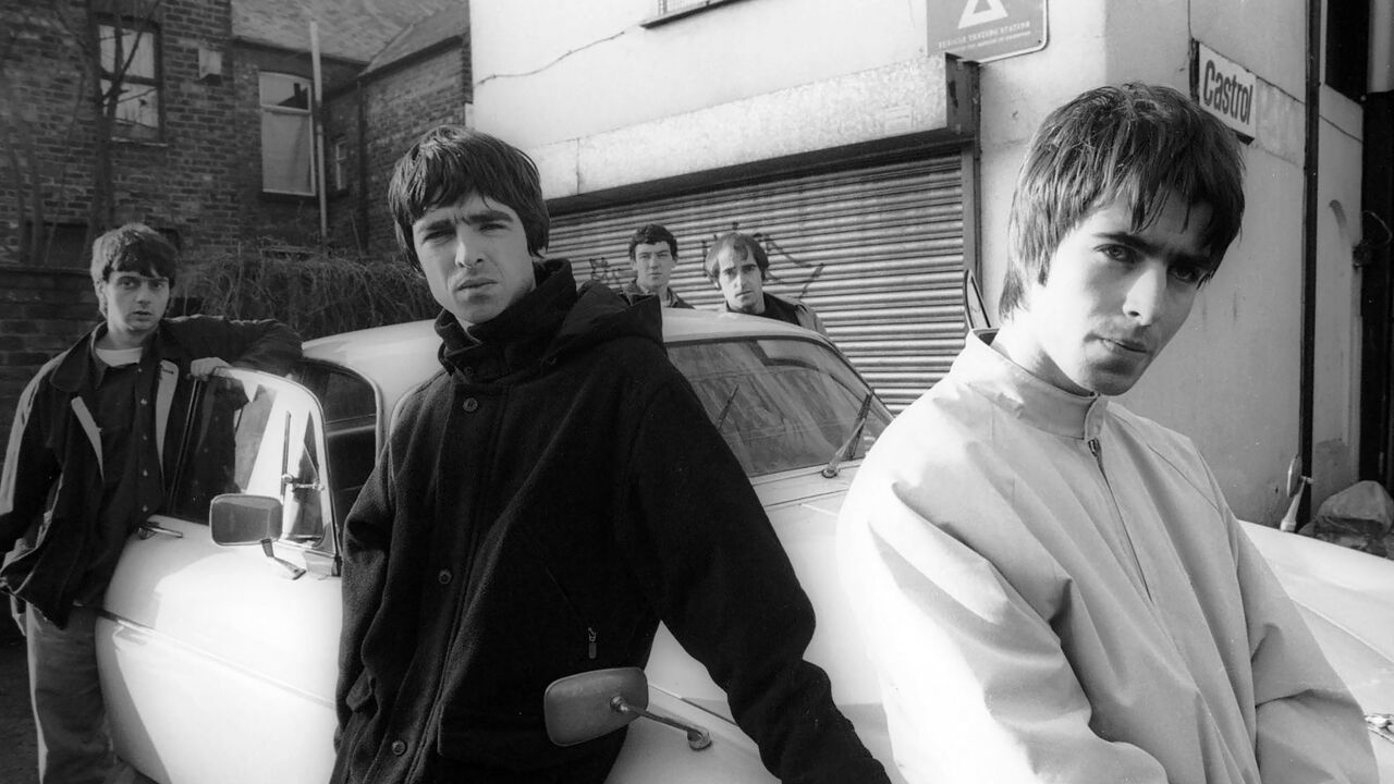 A black and white group portrait of British rock band Oasis leaning against a Jaguar XJ6 car in Withington, Manchester, UK in November 1993.