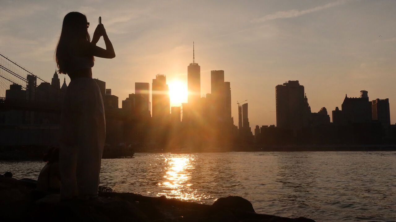  woman photographs the sun setting behind the skyline of lower Manhattan and One World Trade Center from Brooklyn Bridge Park next to the East River in New York City, USA.