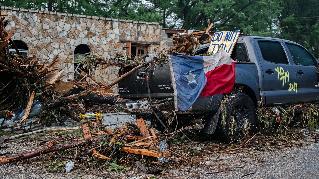 A destroyed SUV draped in a Texas State flag sits next to the road after flash floodingy